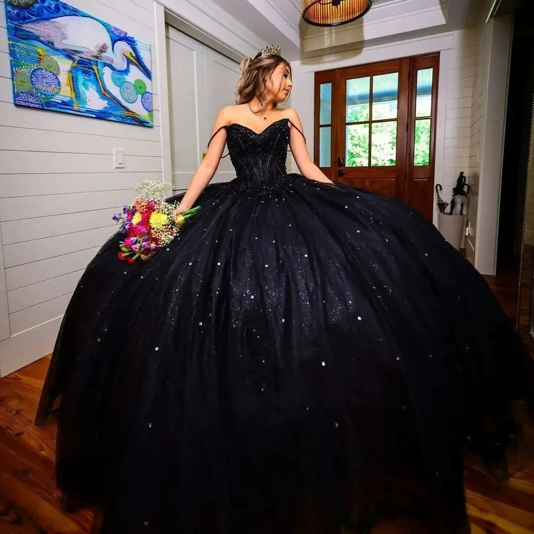 A woman wearing a dramatic black ball gown with a sweetheart neckline, off-the-shoulder straps, and a voluminous sparkly skirt, holding a colorful bouquet in an indoor room with wood floors and a door.