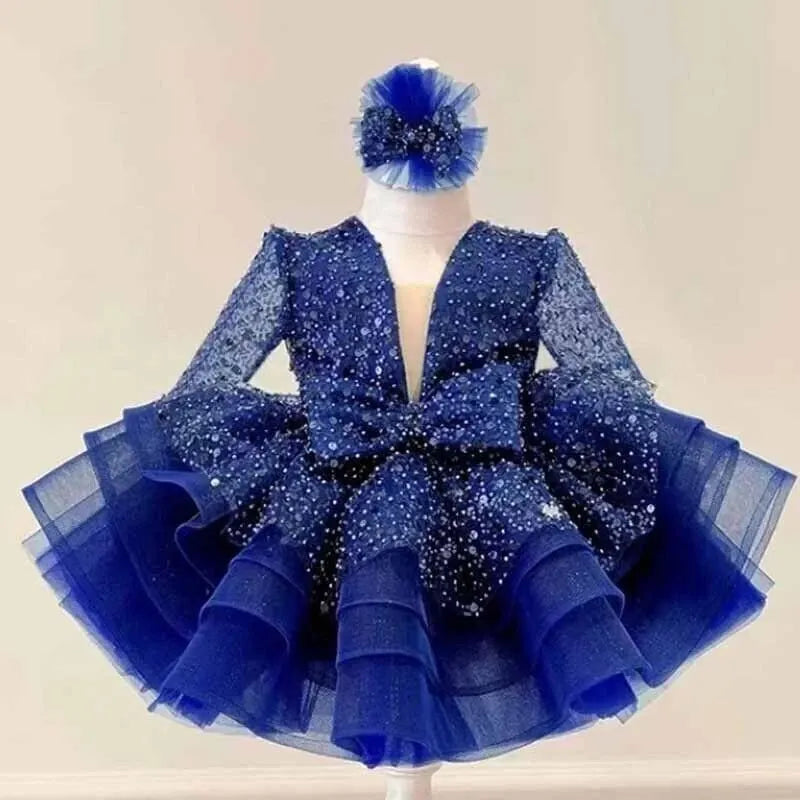A royal blue toddler girl's party dress with a matching headband, The dress features a sparkling sequined bodice, long sheer sleeves, a large central bow, and a tiered tulle skirt.