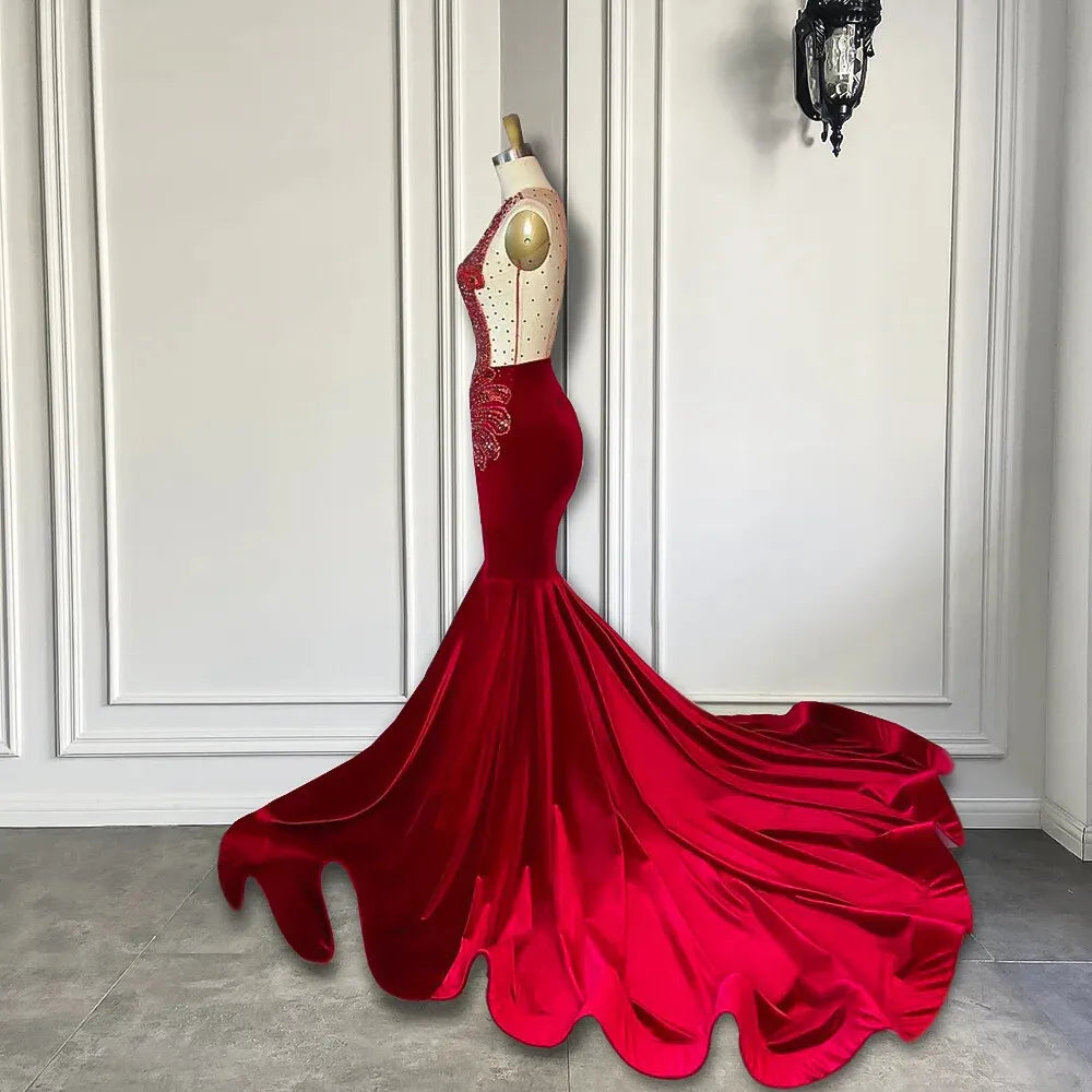 A red formal gown features a sheer lace bodice with gold embroidery, a fitted silhouette, and a dramatic flared satin mermaid skirt with a long train.
