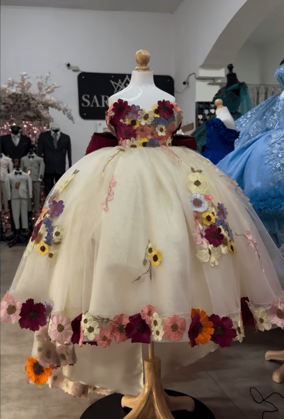 A formal ball gown with a cream tulle skirt with colorful floral appliqués and a matching cream bodice decorated with multicolored flowers.