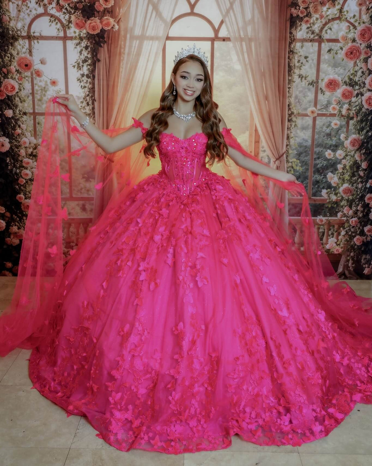 hot pink off-the-shoulder ball gown quince dress with 3D floral applique and a matching sheer pink veil, standing in front of a decorative pink floral backdrop.