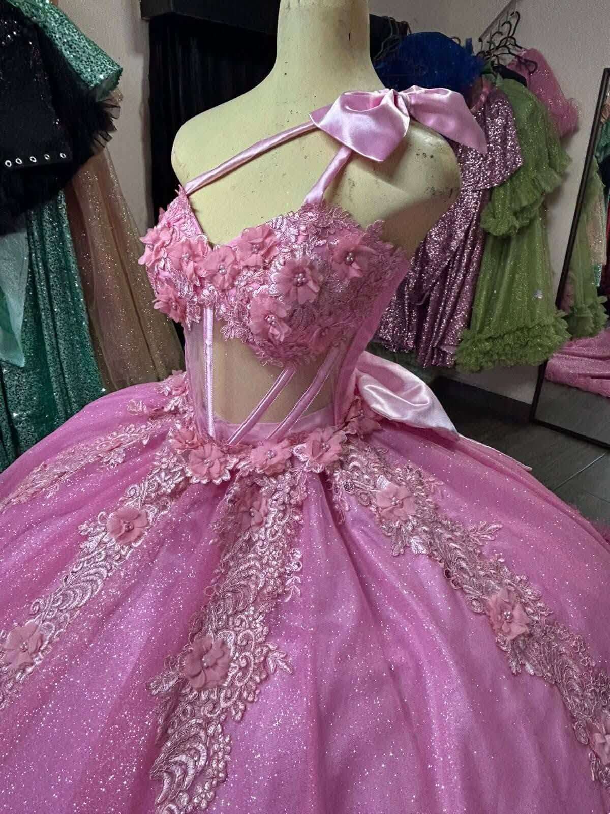 pink ball gown quinceañera dress displayed on a mannequin torso in a retail setting. The dress features a sweetheart neckline with a large satin bow, floral applique, lace detailing, and a voluminous tulle skirt.